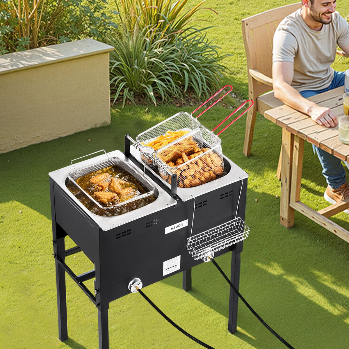 The fryer set up on a lawn with fried chicken in one pan and battered food in the other, and a man sitting at a picnic table behind it