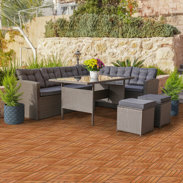 A gray wicker corner sofa, dining table and stools standing on a floor of the tiles, with a stone wall and planting behind