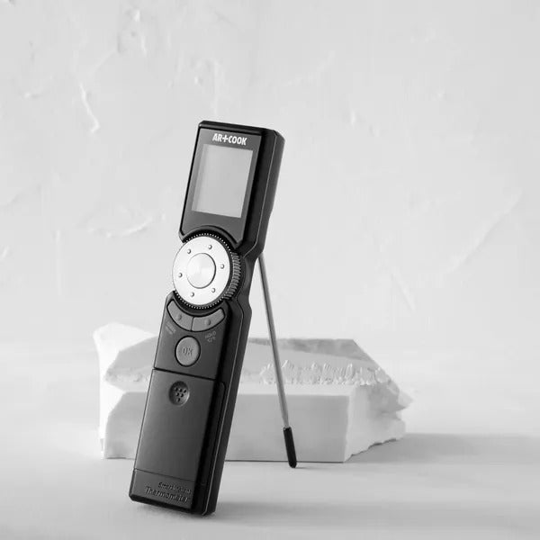 Black digital meat thermometer standing upright on a pale surface with its thin probe folded out to the side.