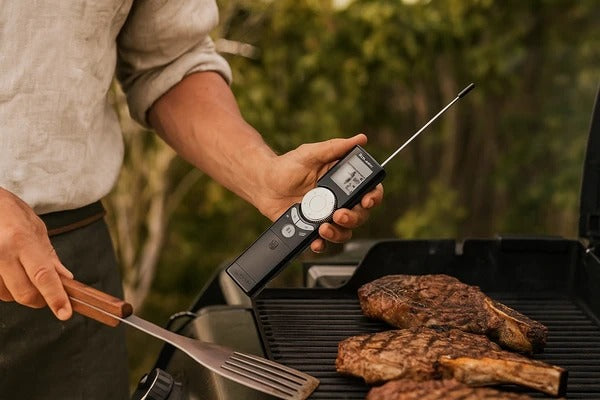 A cook holding the thermometer in one hand and a spatula in the other beside steaks on an outdoor grill.
