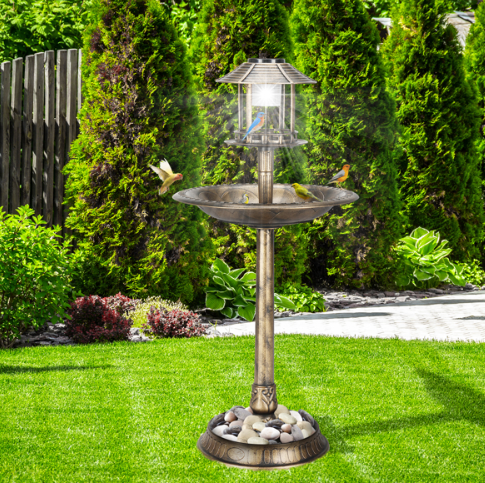 The bird bath on a mown lawn in front of a tall evergreen hedge, with birds at the basin and white stones filling the base